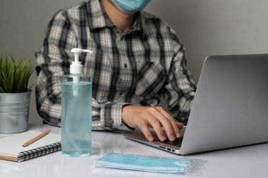 Coronavirus hastalığı veya COVID-19 koruma kavramı - Genç adam, Corona virüsüne karşı korunmak için masadaki dizüstü bilgisayarı kullanırken koruyucu ve temizlik ekipmanlarıyla evde çalışıyor.