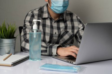 Coronavirus hastalığı veya COVID-19 koruma kavramı - Genç adam, Corona virüsüne karşı korunmak için masadaki dizüstü bilgisayarı kullanırken koruyucu ve temizlik ekipmanlarıyla evde çalışıyor.