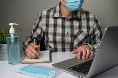 Coronavirus hastalığı veya COVID-19 koruma kavramı - Genç adam, Corona virüsüne karşı korunmak için masadaki dizüstü bilgisayarı kullanırken koruyucu ve temizlik ekipmanlarıyla evde çalışıyor.