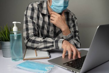 Coronavirus hastalığı veya COVID-19 koruma kavramı - Genç adam, Corona virüsüne karşı korunmak için masadaki dizüstü bilgisayarı kullanırken koruyucu ve temizlik ekipmanlarıyla evde çalışıyor.