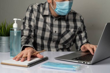Coronavirus hastalığı veya COVID-19 koruma kavramı - Genç adam, Corona virüsüne karşı korunmak için masadaki dizüstü bilgisayarı kullanırken koruyucu ve temizlik ekipmanlarıyla evde çalışıyor.